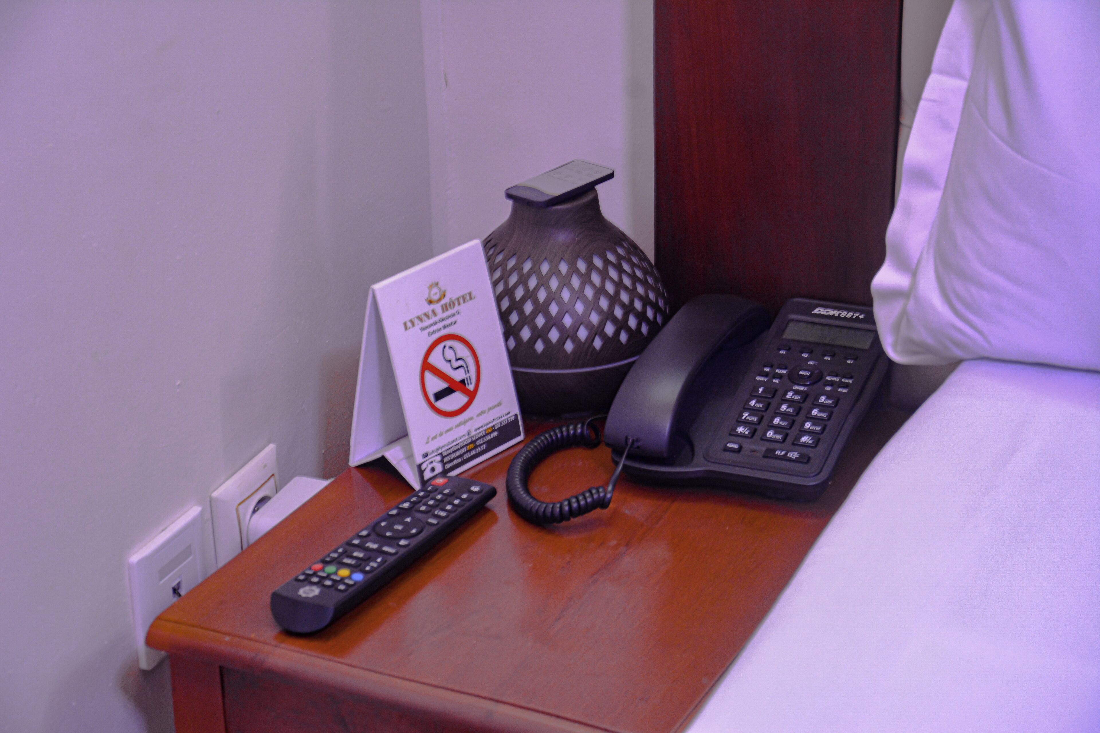 A hotel room with a bedside table that includes a remote control, a telephone, and a sign indicating no smoking.