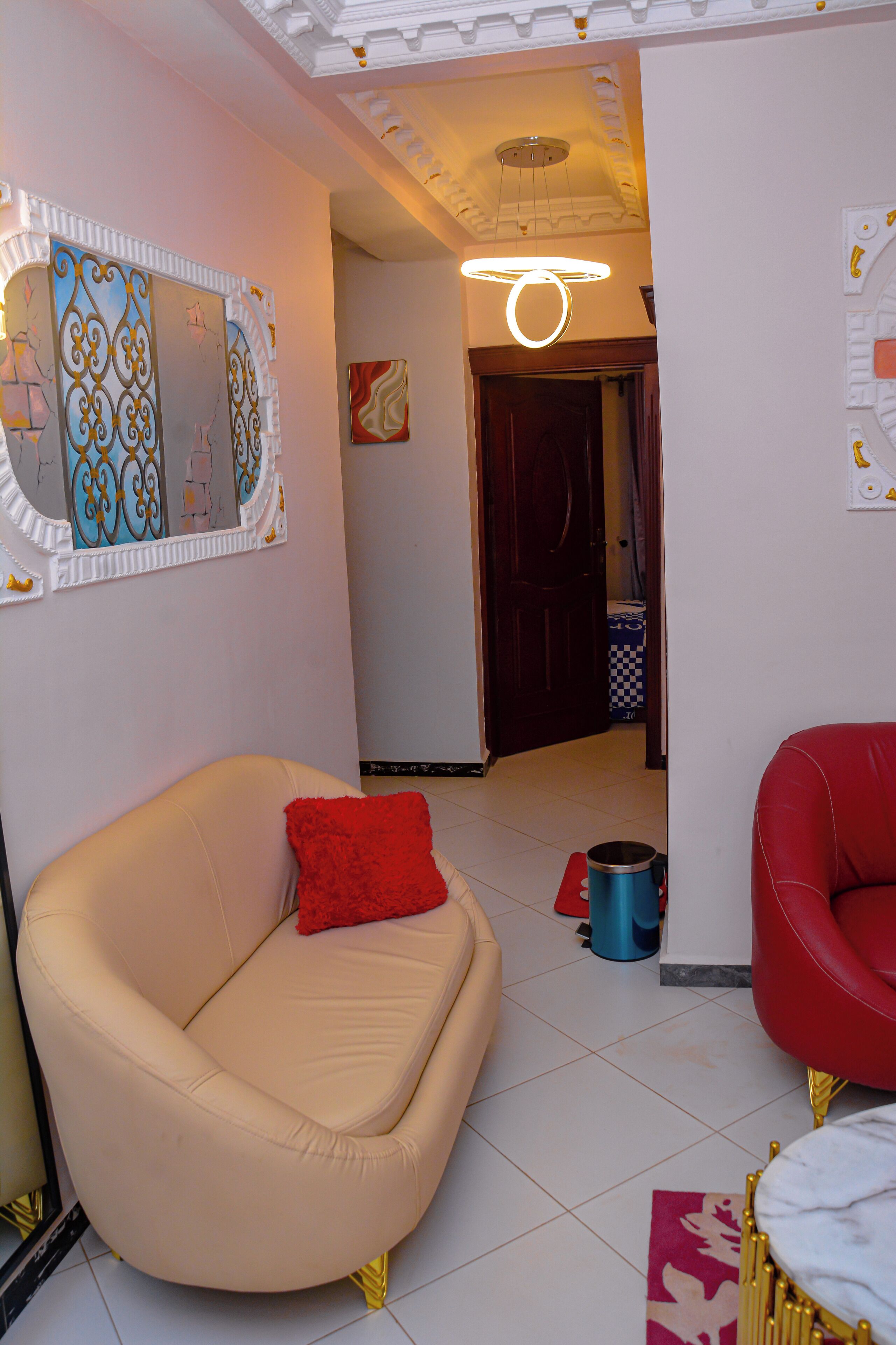 Un soggiorno con un divano beige, una poltrona rossa e un cuscino decorativo rosso. Si nota un lampadario a soffitto e uno specchio appeso alla parete.