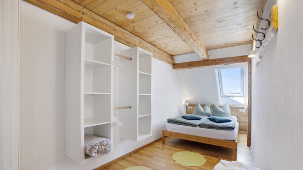 Una camera da letto con soffitto in legno, un letto con cuscini, un armadio bianco e una finestra.