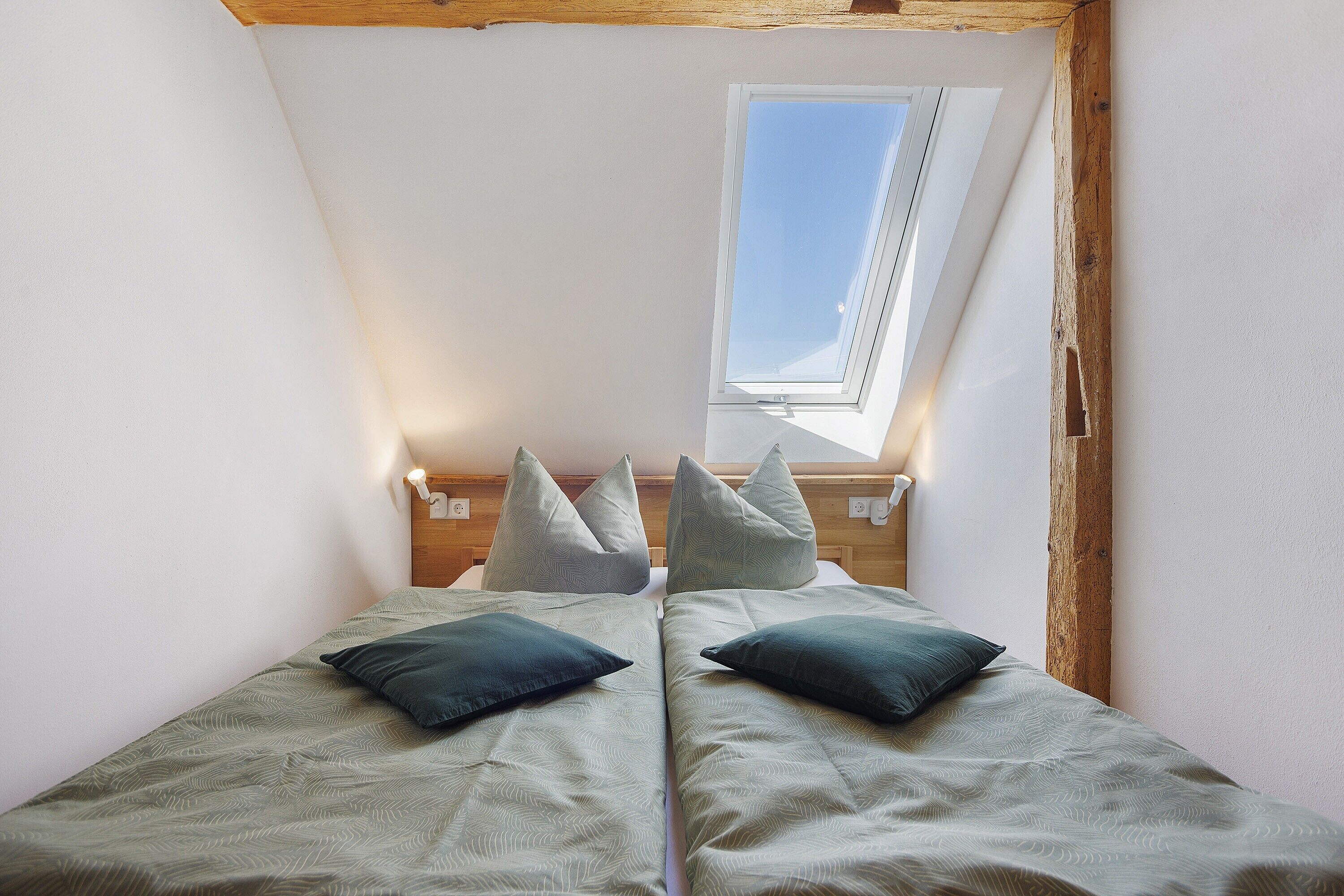 A room with a sloped ceiling, a skylight, two beds with pillows, and wooden beams.