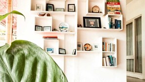 A room with a white bookshelf filled with books and decorative items, a large green plant in the foreground, and a patterned floor. - Lacasa Senggigi By Royal Amreetha (Senggigi)