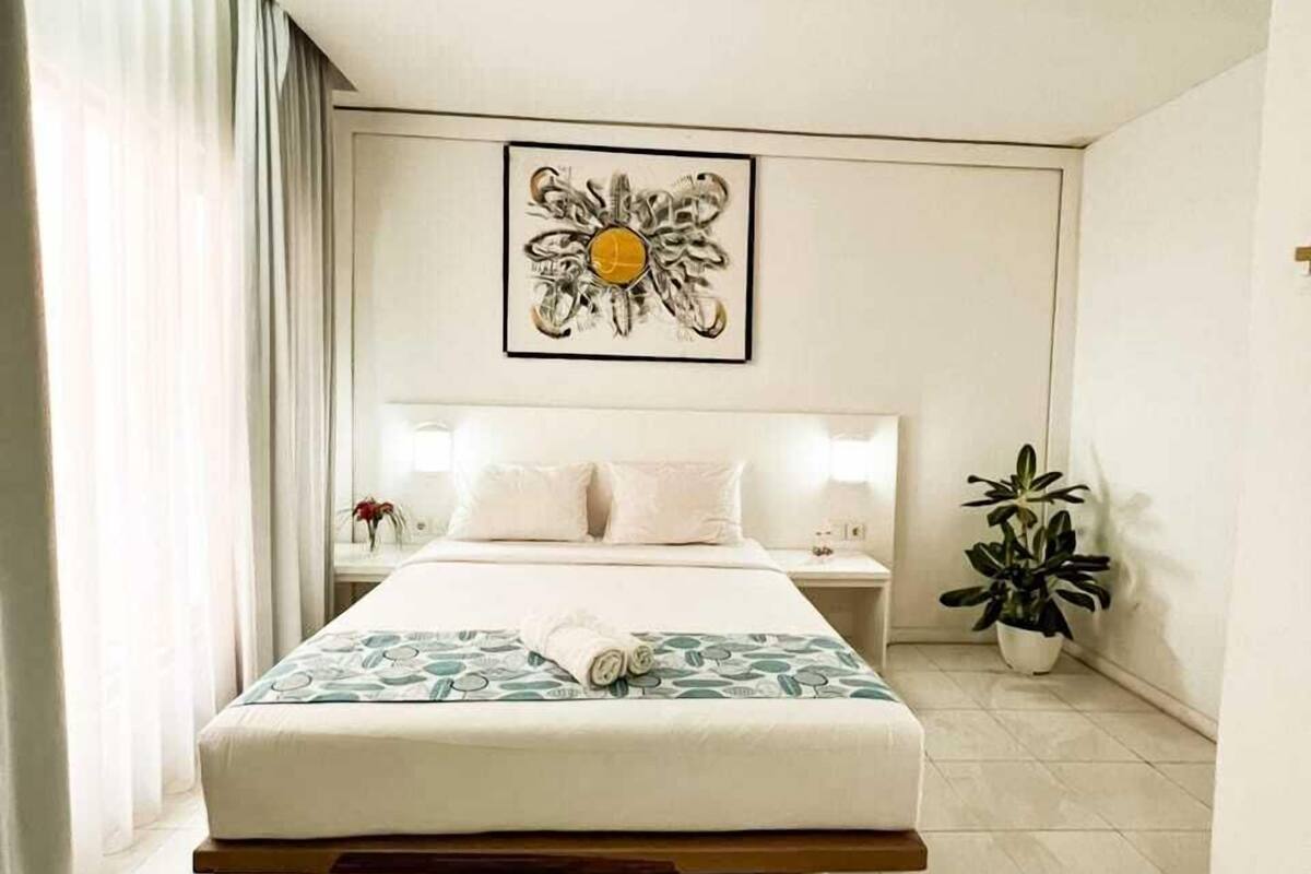 Une chambre d’hôtel avec un lit, une table de chevet, une plante en pot et un tableau encadré au mur.