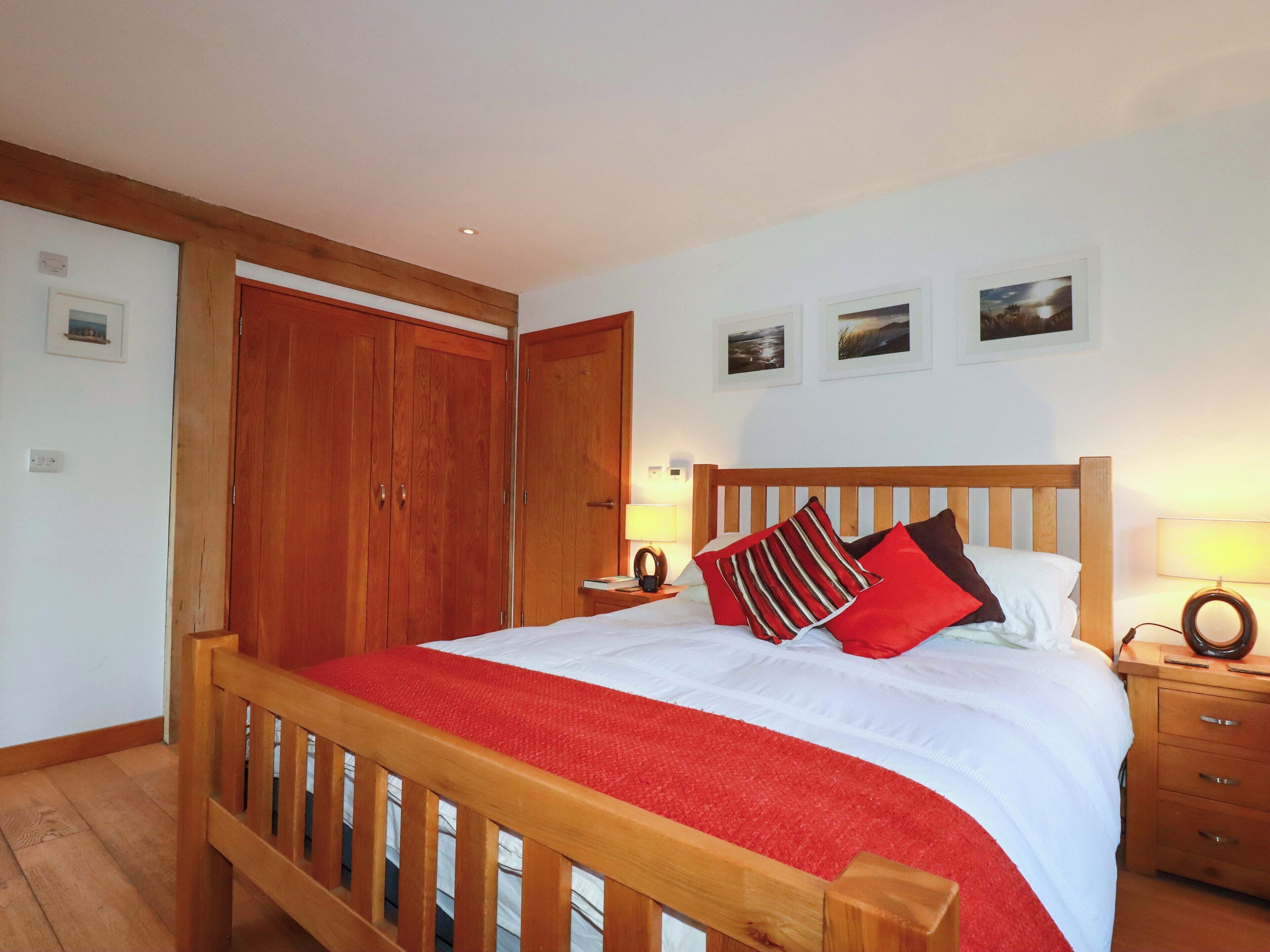 A bedroom with a wooden bed frame, a red bedspread, and a nightstand with a lamp. There are two wooden wardrobes and a door to the left.