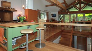A spacious kitchen with wooden beams, a large island with stools, and a view of the outdoors through large windows. - Kerslake House (Torpoint)