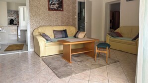 Sala de estar com um sofá bege, mesa de centro de madeira e uma cadeira pequena. Há um quadro na parede e uma porta que dá para outro cômodo.