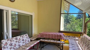 Uma área de pátio coberta com mesa, bancos, grandes janelas e vista para o verde.