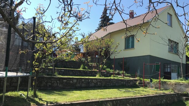 Um edifício de dois andares com telhado inclinado, cercado por um muro de pedra e uma área gramada com árvores.