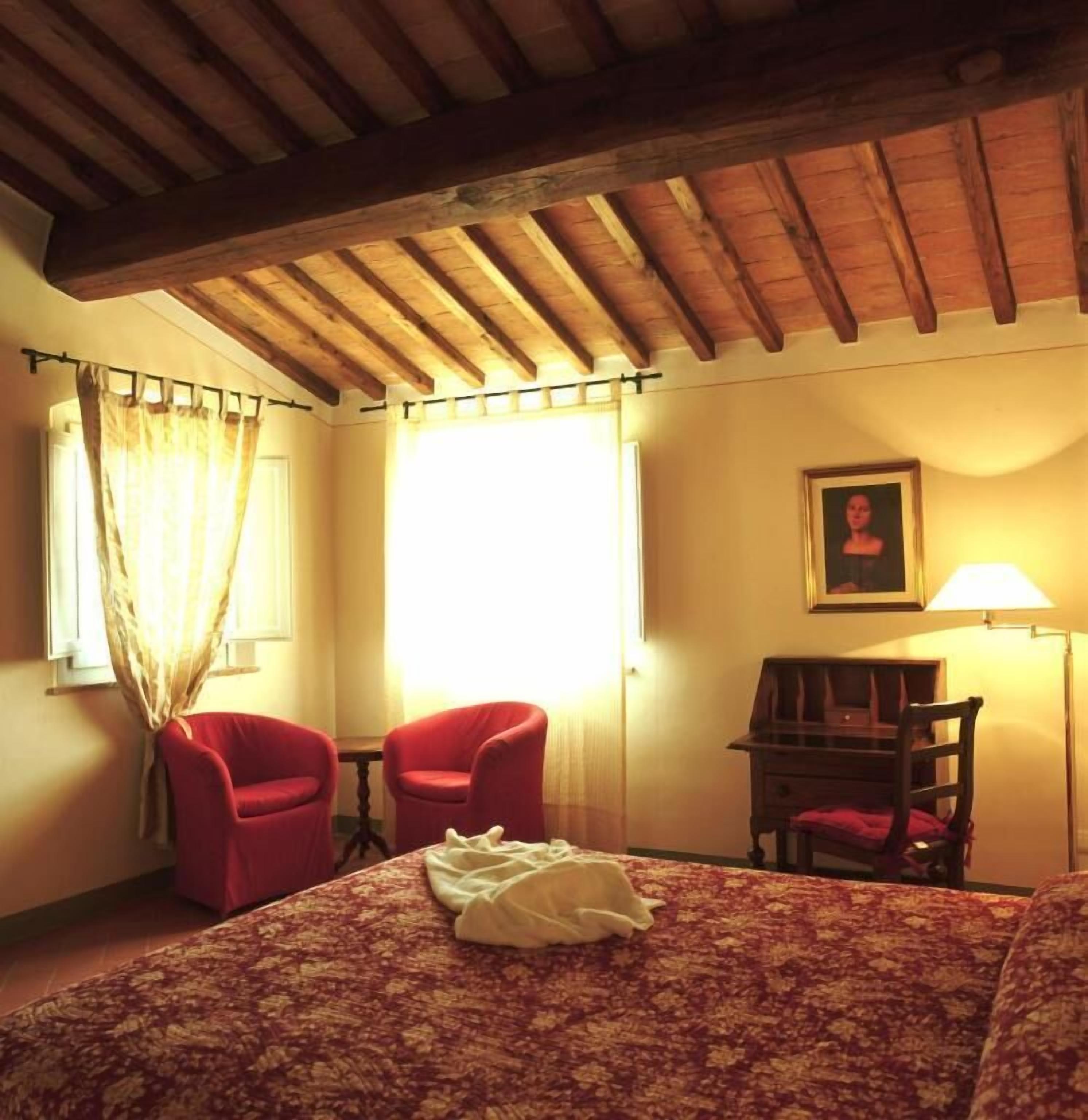 A hotel room with a wooden ceiling, a bed with a floral bedspread, two red chairs, a desk with a lamp, and a framed picture on the wall.