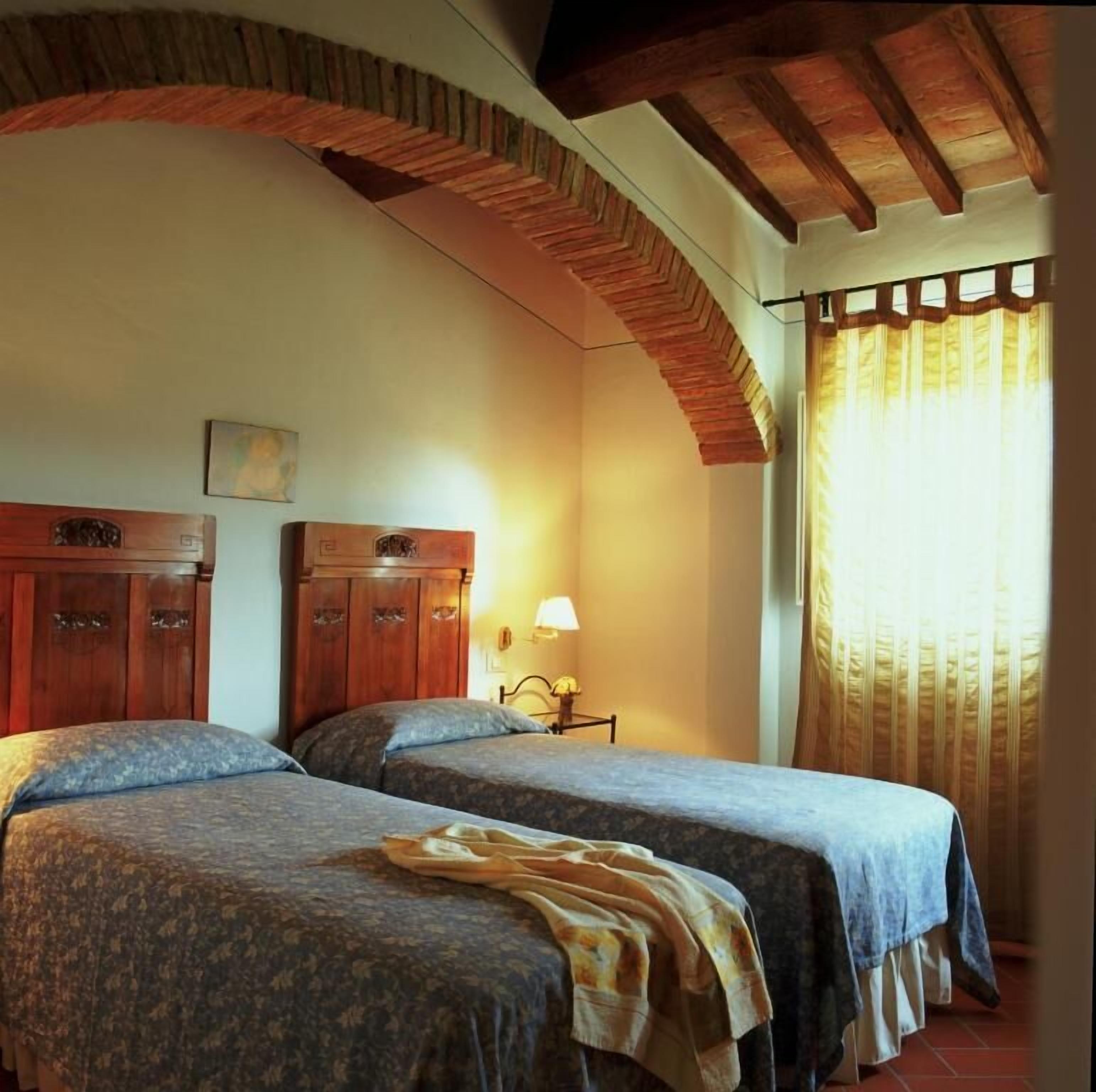 Un dormitorio con dos camas, cabeceros de madera, una mesita de noche con lámpara y una ventana con cortinas.