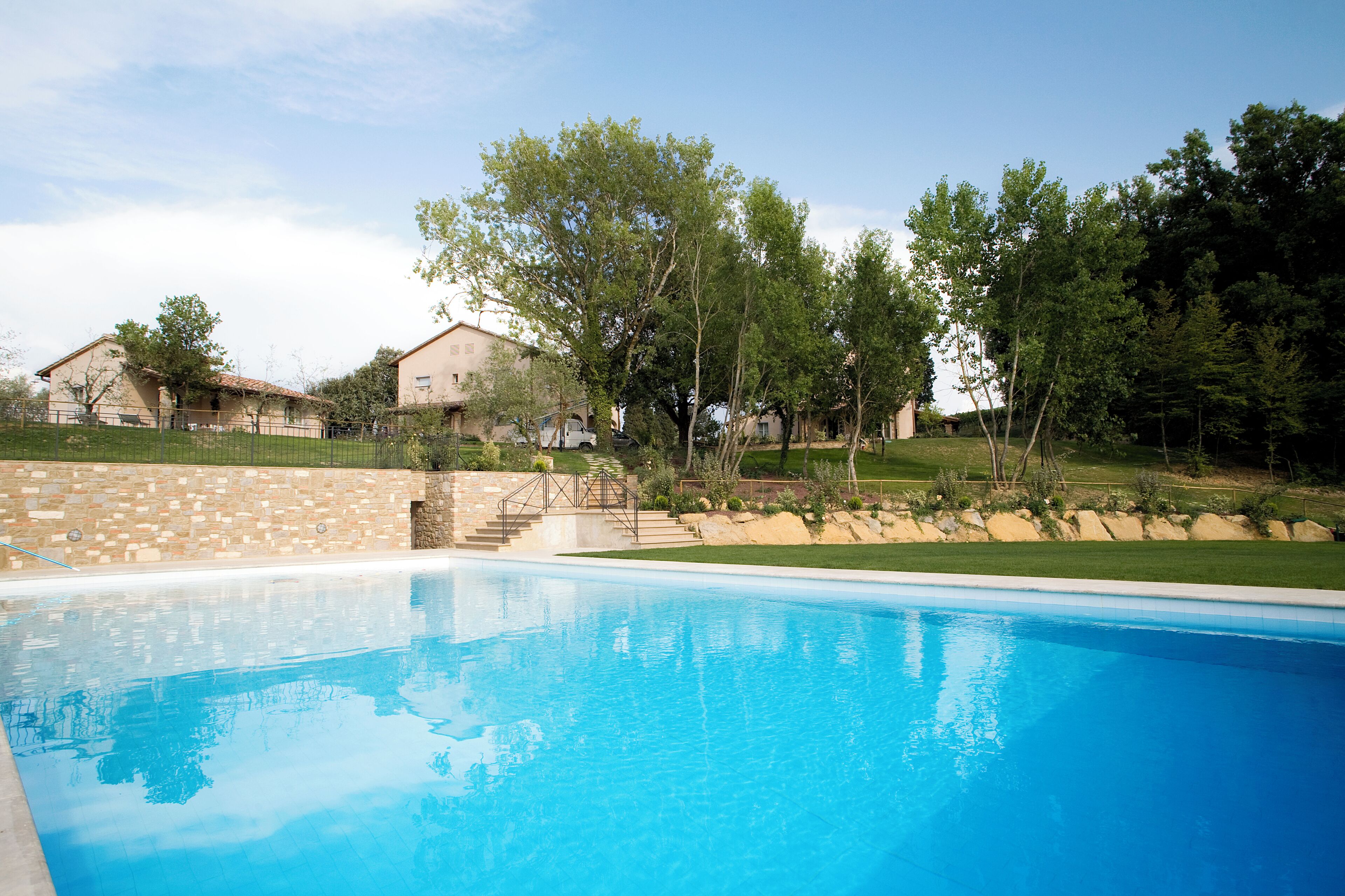 Una piscina con un muro de piedra y un área verde con árboles y casas al fondo.