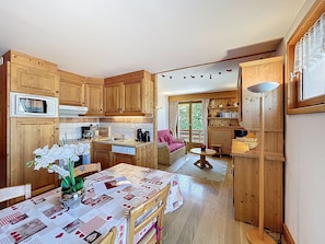 A cozy kitchen with wooden cabinets, a dining table with a quilted tablecloth, and a view of the outdoors through a sliding glass door. - 3-Room Apartment with Parking and Mont Blanc Views (Combloux)