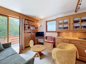 Een gezellige woonkamer met een bank, fauteuil, houten kasten, een televisie en een balkon met uitzicht op bomen.