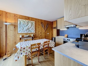A cozy kitchen with wooden walls, a dining table with a fruit-patterned tablecloth, and a modern stove. - 3-Room Apartment with Parking and Mont Blanc Views (Combloux)