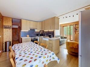 A compact kitchen with wooden cabinets, a central island, and a dining area with a table covered in a patterned tablecloth. - 3-Room Apartment with Parking and Mont Blanc Views (Combloux)