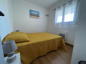 Una habitación pequeña con cama individual, colcha amarilla, un cuadro con barcos en la pared y una ventana con cortinas.
