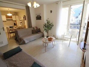A spacious living area with a sofa, a dining table with a vase and a basket, a balcony with a chair, and a kitchen in the background. - Central Luchon Apartment - 2 Rooms - 4 Pers - Pets OK (Bagnères-de-Luchon)