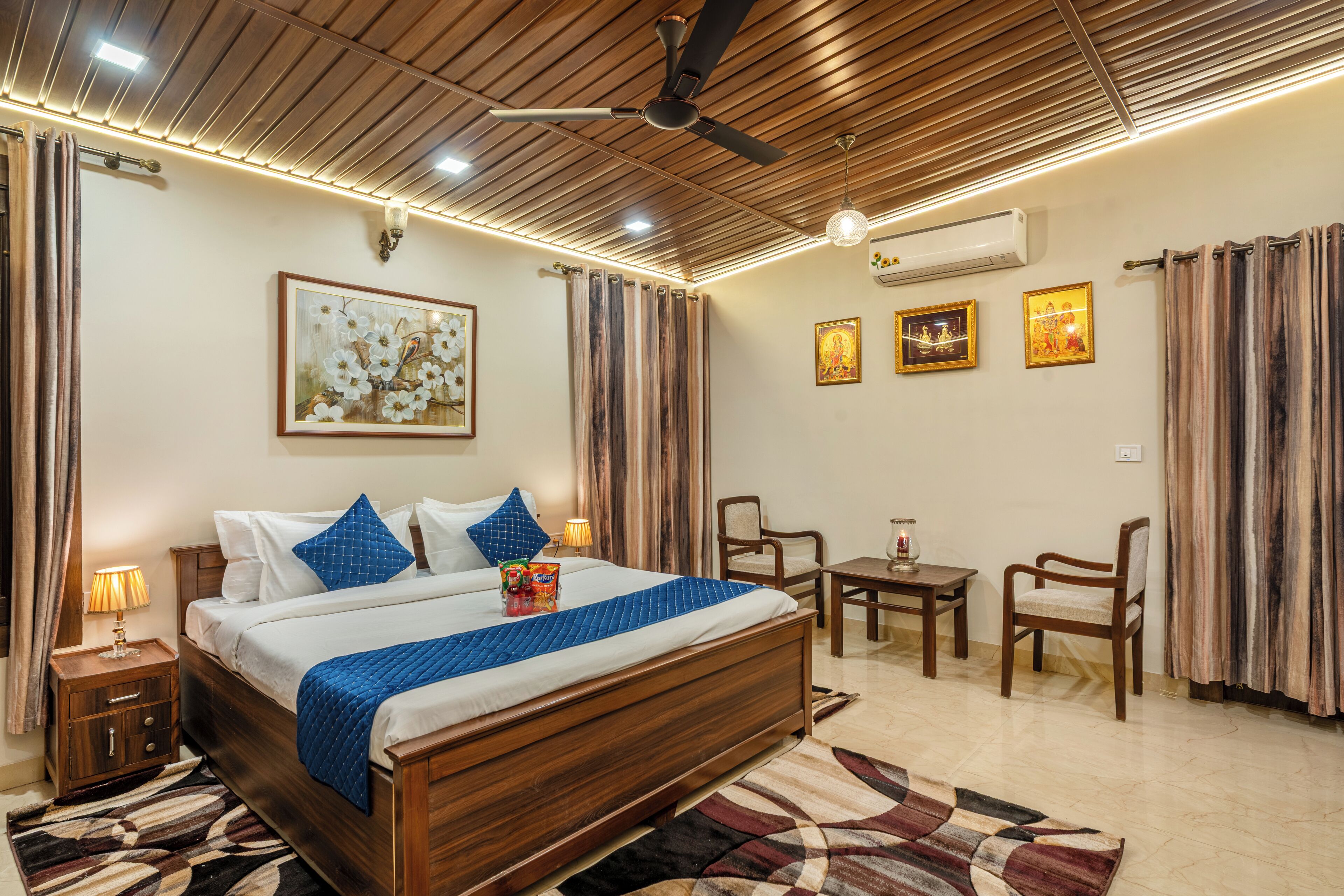 A well-lit bedroom with a wooden ceiling, a bed with blue bedding, a ceiling fan, and a small table with chairs.