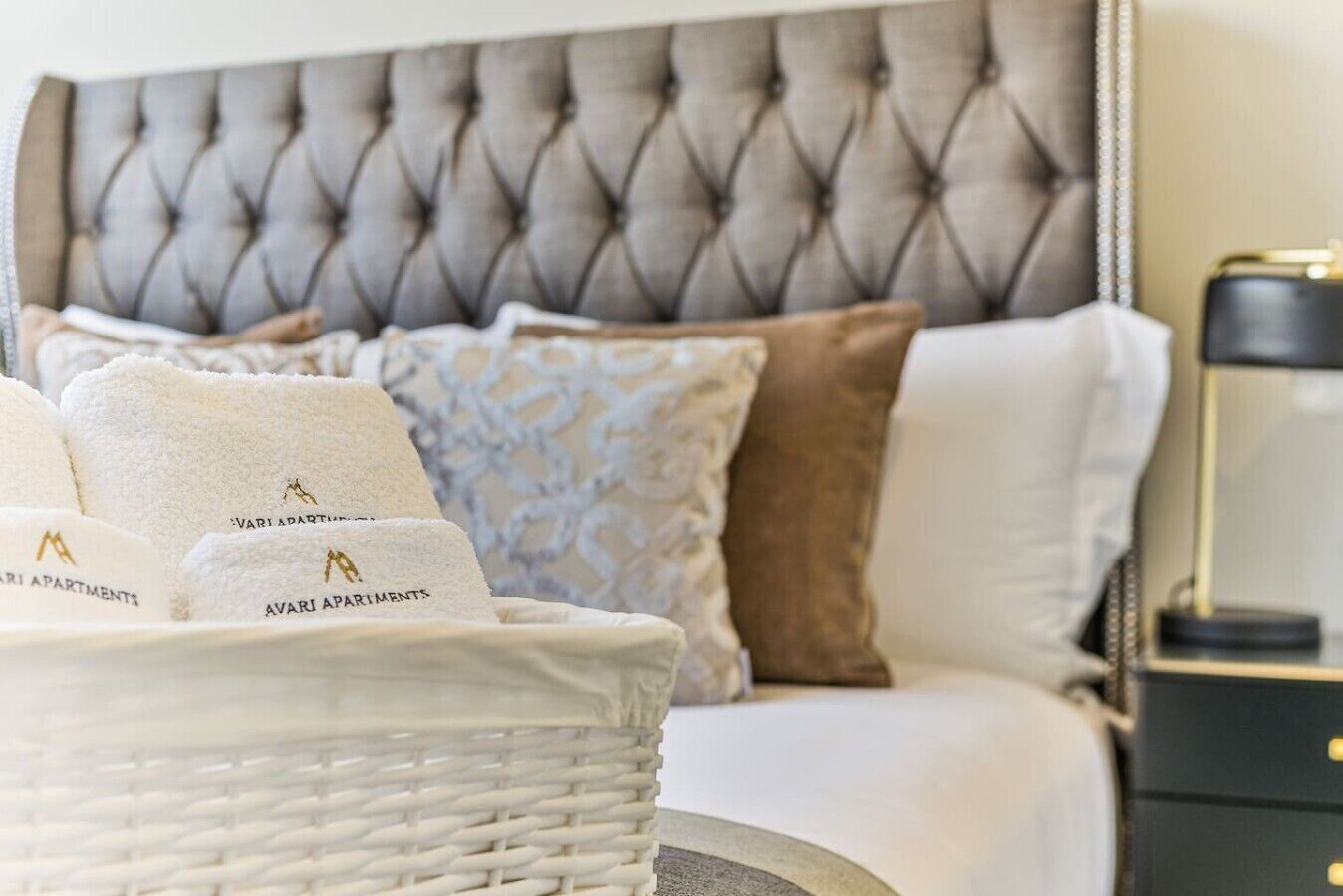 A bed with a tufted headboard, a basket of towels, and a bedside table with a lamp.