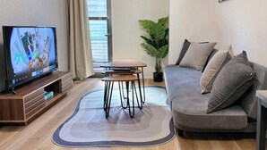 Ett vardagsrum med en platt-TV, en grå soffa, ett litet träbord och en krukväxt.