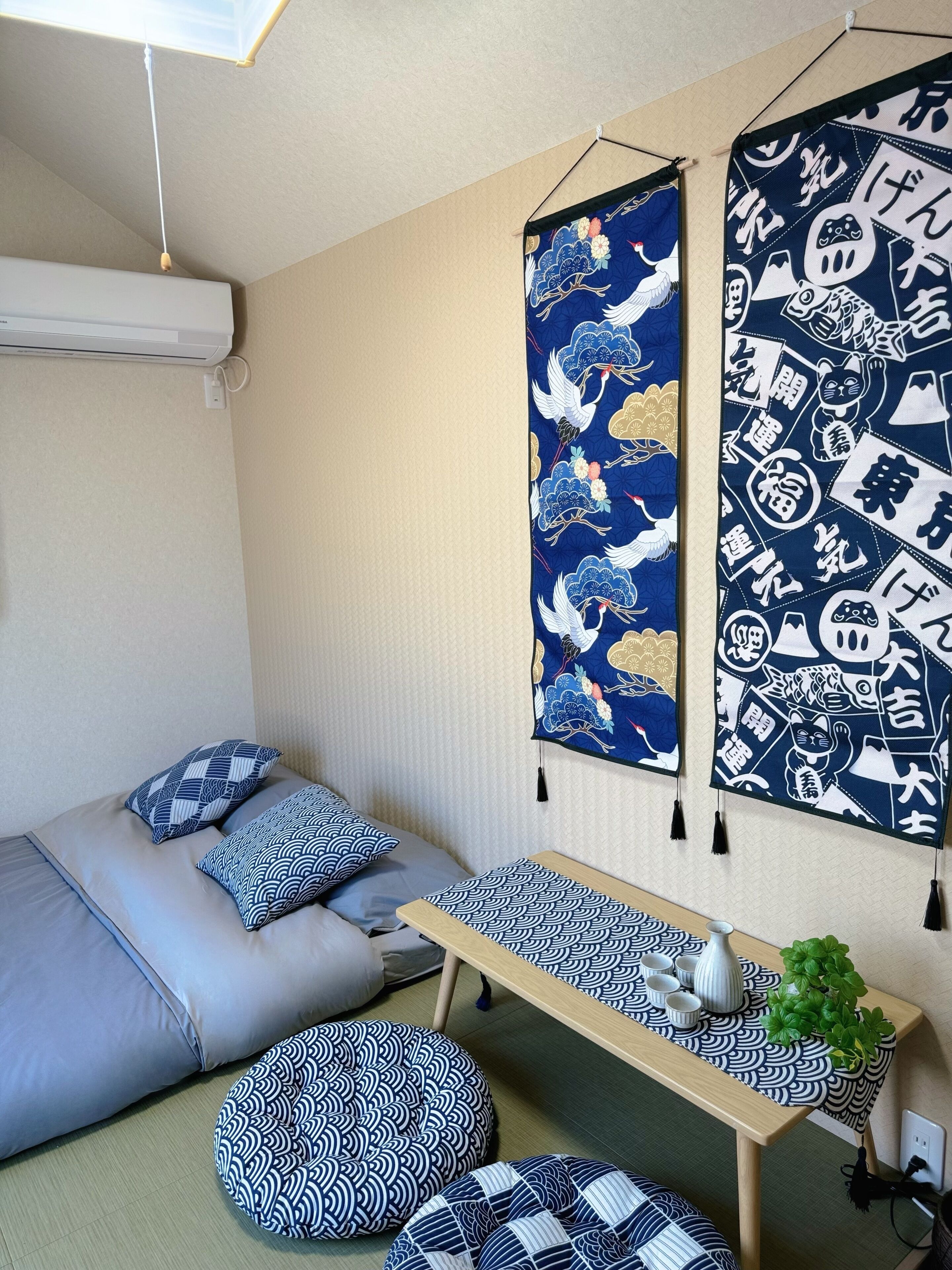 A room with a bed, pillows, a table with a plant, and two wall hangings with traditional patterns.