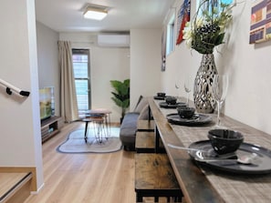 Een moderne eethoek met een lange houten tafel gedekt voor vier personen, een bloemenvaas en een televisie op de achtergrond.