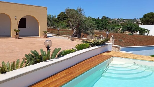A pool area with a wooden deck, a white building with arches, and a view of a hillside with trees. - Apartment 'Appartamento Cosimo' with Sea View, Wi-Fi and Air Conditioning (Marina di Pescoluse)