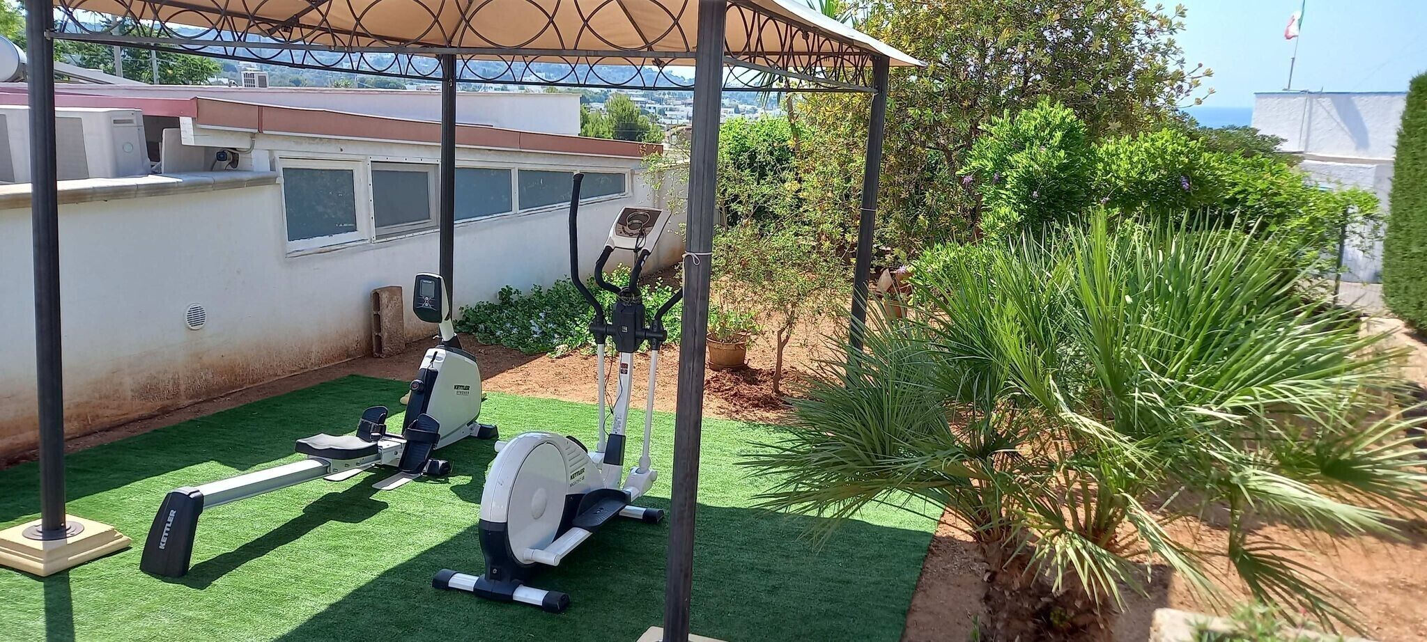 An outdoor gym area with stationary bikes and a treadmill, surrounded by greenery and a building in the background.