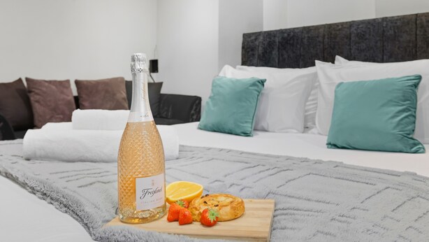 Ein ordentlich bezogenes Bett mit weißen und türkisfarbenen Kissen, eine Flasche Freixenet Champagner und ein Schneidebrett mit Erdbeeren und einem Gebäckstück.