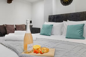 Un lit bien fait, agrémenté d’oreillers blancs et turquoise, une bouteille de champagne Freixenet et une planche à découper avec des fraises et une pâtisserie.