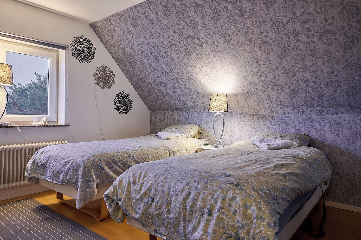 A bedroom with two beds, a radiator, a lamp, and a window with a view of trees.