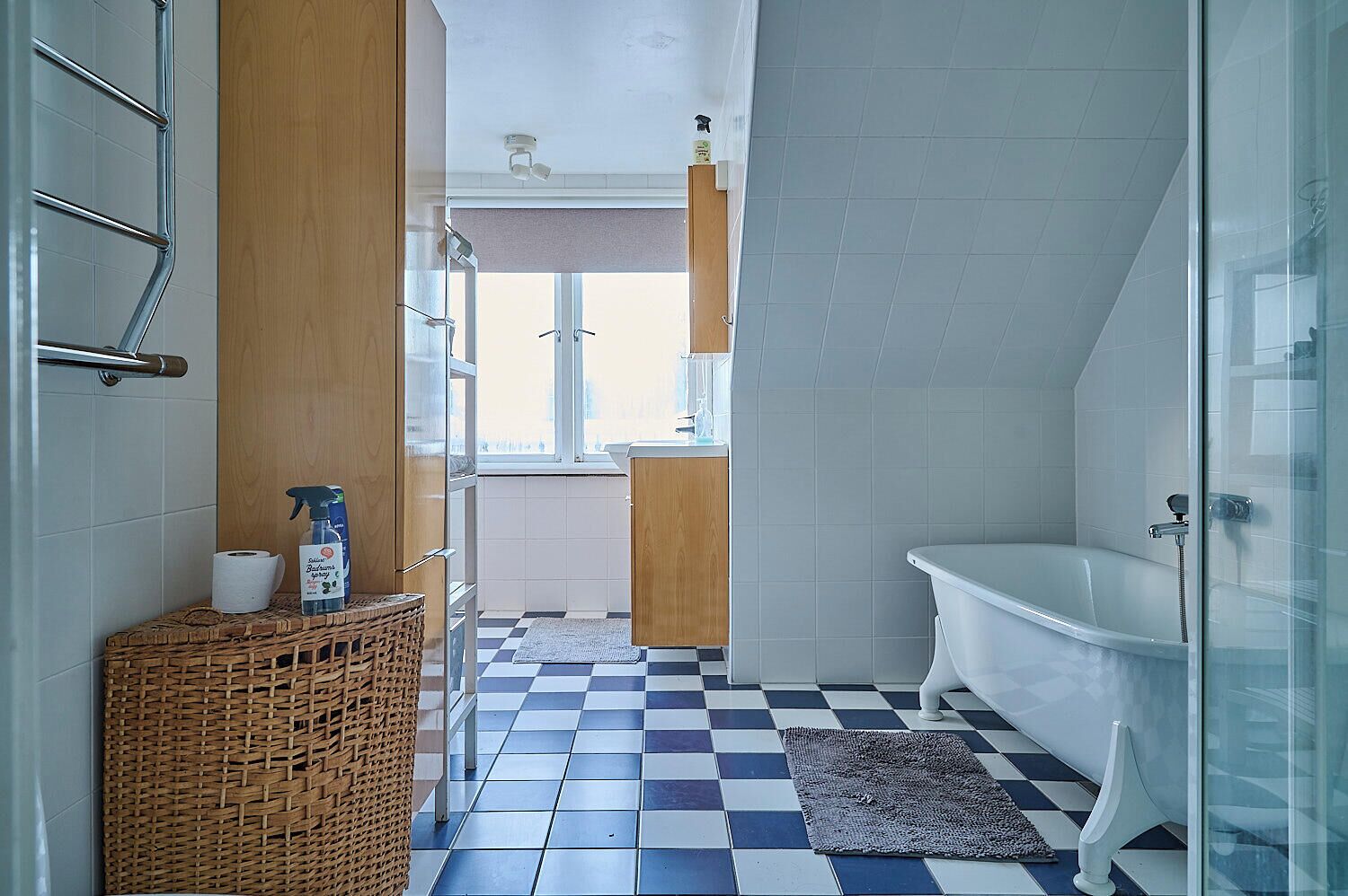 Ein Badezimmer mit Schachbrettfußboden, einer weißen Badewanne und einem Korbwäscher mit Toilettenartikeln.