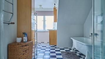 Ein Badezimmer mit Schachbrettfußboden, einer weißen Badewanne und einem Korbwäscher mit Toilettenartikeln.