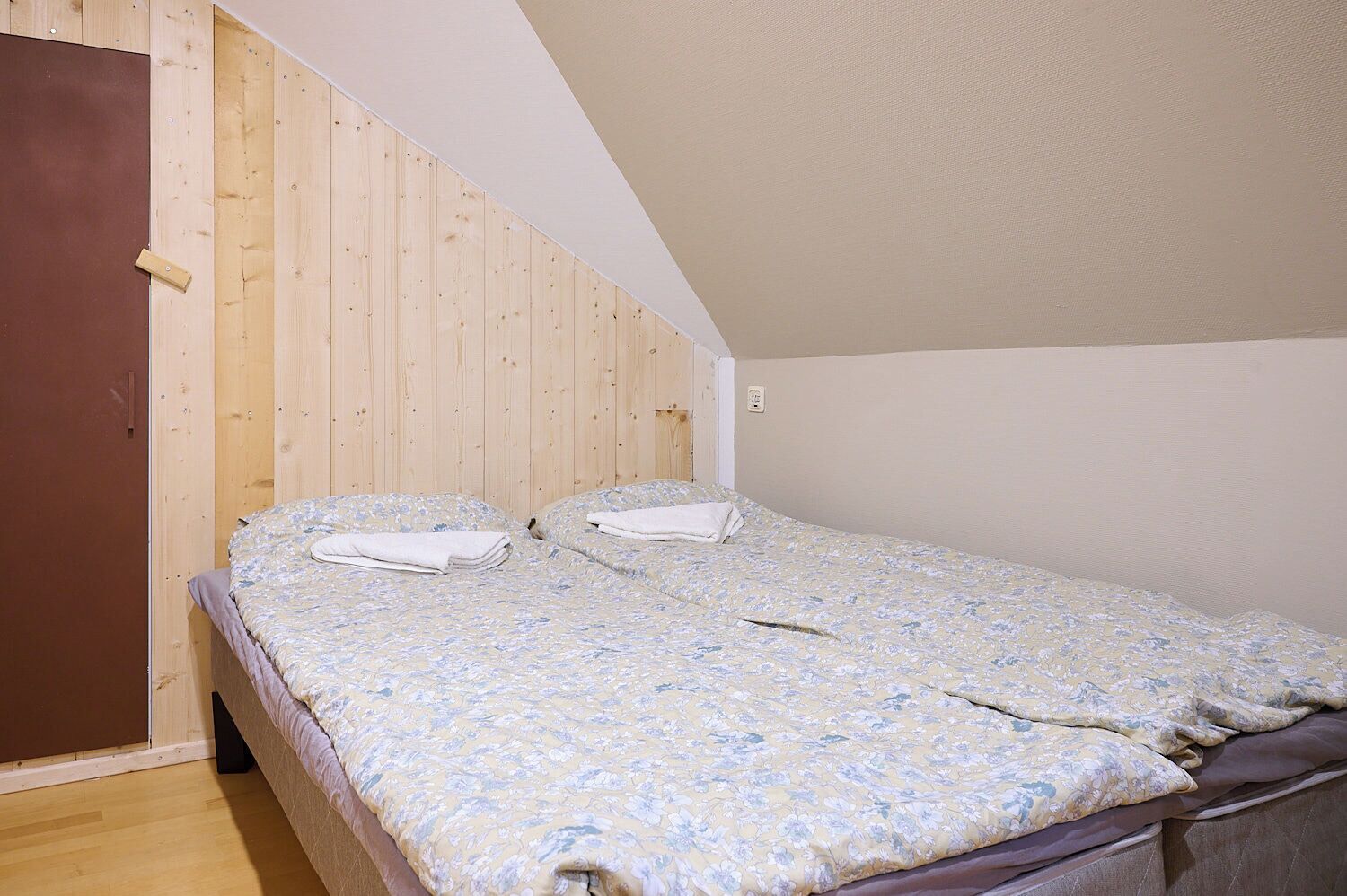 Uma cama de solteiro com colcha floral num quarto com paredes de madeira e teto inclinado.