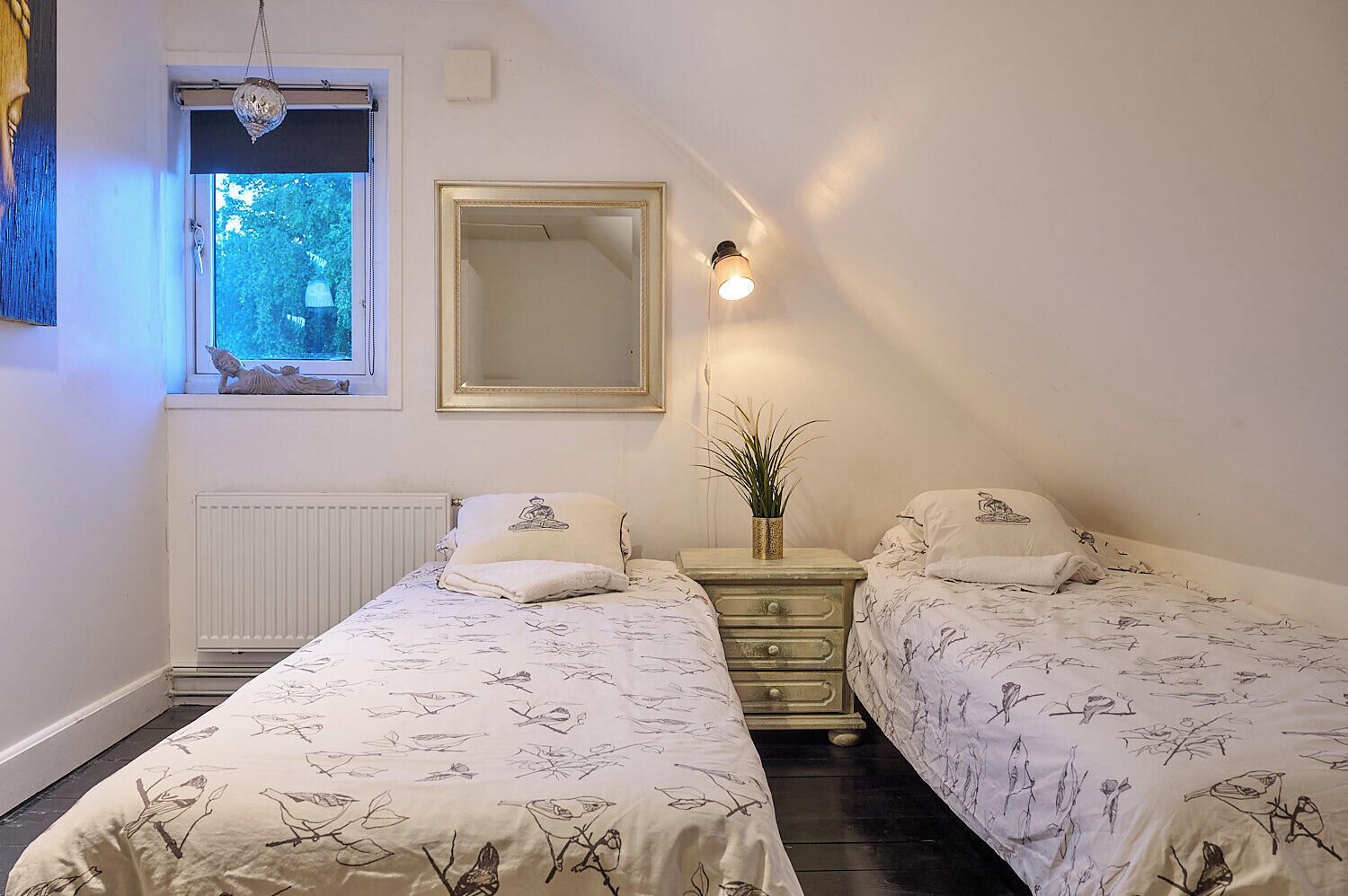 A bedroom with two beds, a dresser, a mirror, a radiator, and a window with a view of trees.
