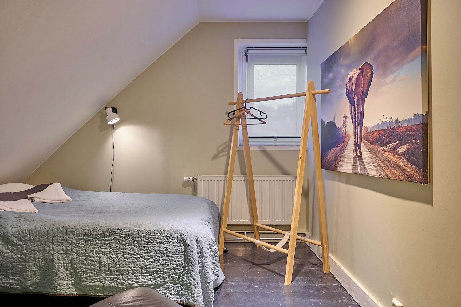 A bedroom with a bed, a wooden hanger rack, a radiator, a window, and a painting of an elephant on the wall.