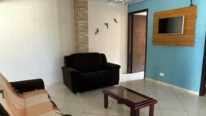 A living room with a sofa, a coffee table, a black armchair, a TV mounted on the wall, and a ceiling light.