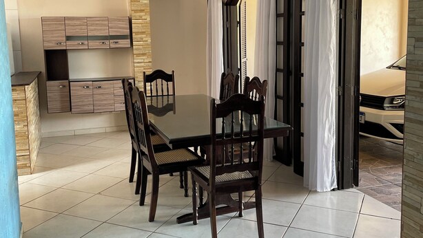 A dining area with a wooden table and chairs, a kitchen in the background, and a view through an open door.