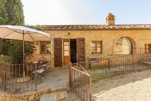 Un bâtiment en pierre avec un toit en tuiles, un espace extérieur aménagé avec une table et des chaises, et une terrasse couverte avec un parasol.