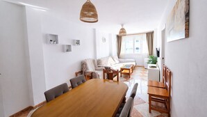 Un'ampia zona giorno con un tavolo da pranzo in legno, divani e un tavolino. Sono presenti due lampade pendenti in vimini e un quadro appeso al muro.