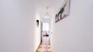 Un corridoio con pareti bianche, pavimento a scacchi, una pianta sospesa, una lampada a sospensione e un quadro appeso al muro.