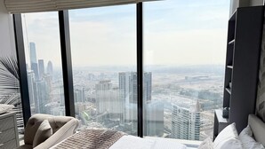 Habitación de hotel moderna con una cama grande, zona de estar y vista panorámica de la ciudad a través de ventanales del suelo al techo.