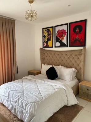 A bedroom with a tufted headboard, a white bedspread, a black pillow, and three framed artworks on the wall. - 3As Properties (Accra)