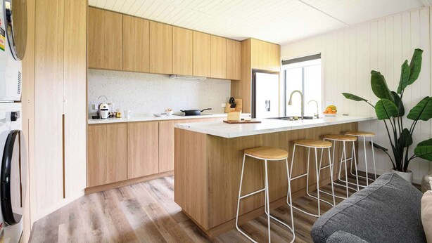A modern kitchen with wooden cabinets, a large island with stools, a sink, and a window. - Wood Duck Daylesford (Daylesford)
