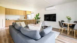 A modern living room with a grey sectional sofa, a flat-screen TV mounted on the wall, and a dining area with a round table and chairs. - Wood Duck Daylesford (Daylesford)