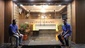 Deux hommes sont assis sur des tabourets dans le hall d’un hôtel, devant un comptoir d’accueil, et une enseigne indique : « PRATAPGRAND HOTEL ».