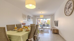 Un comedor con sillas de mimbre, mantel verde, alimentos para el desayuno y un gran reloj de pared.