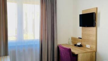 Ein Hotelzimmer mit einem Bett, einem Schreibtisch mit Stuhl, einem an der Wand befestigten Fernseher und einem Fenster mit Vorhängen.