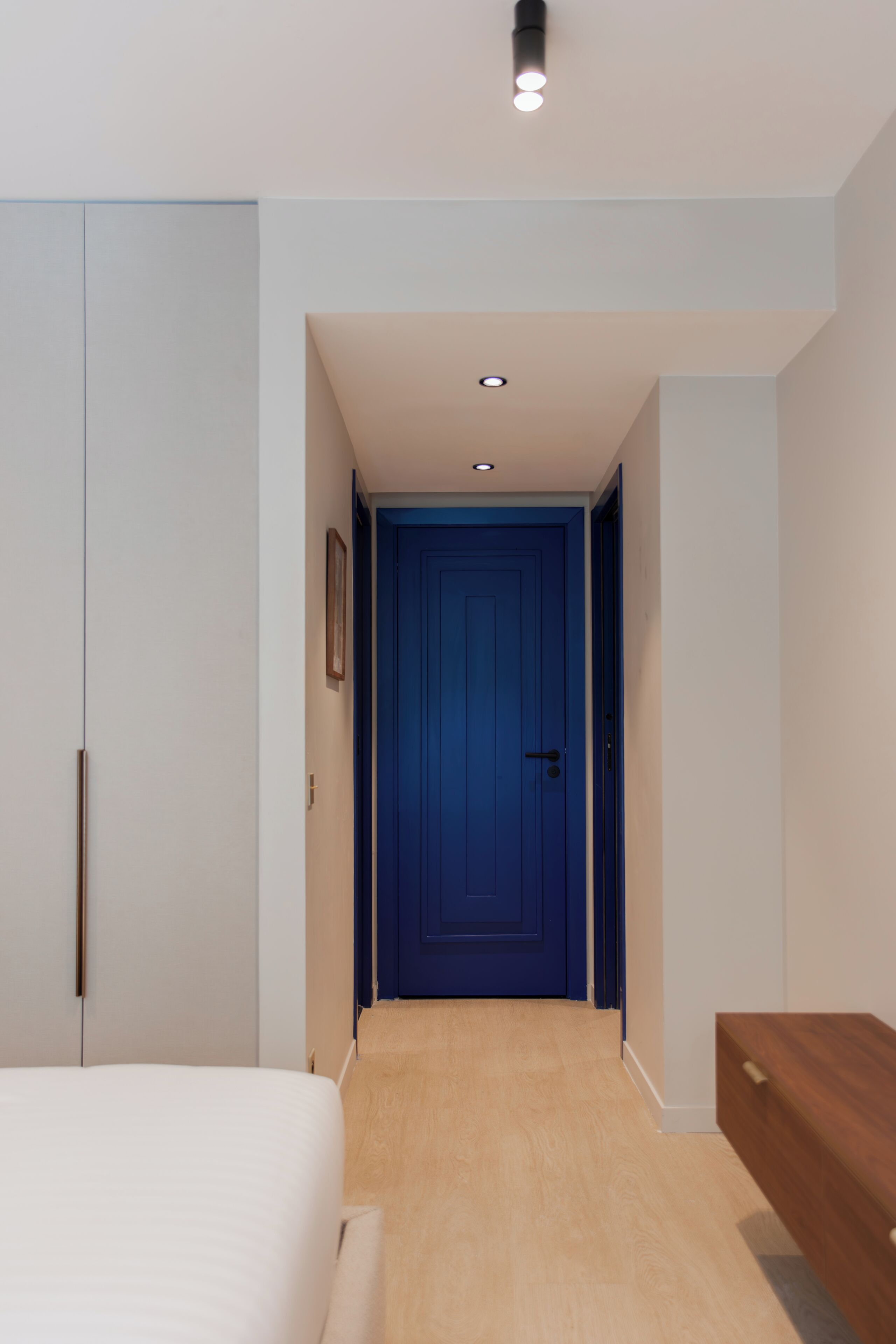 A narrow hallway with a blue door at the end, a bed with a white headboard, and a wooden bench.