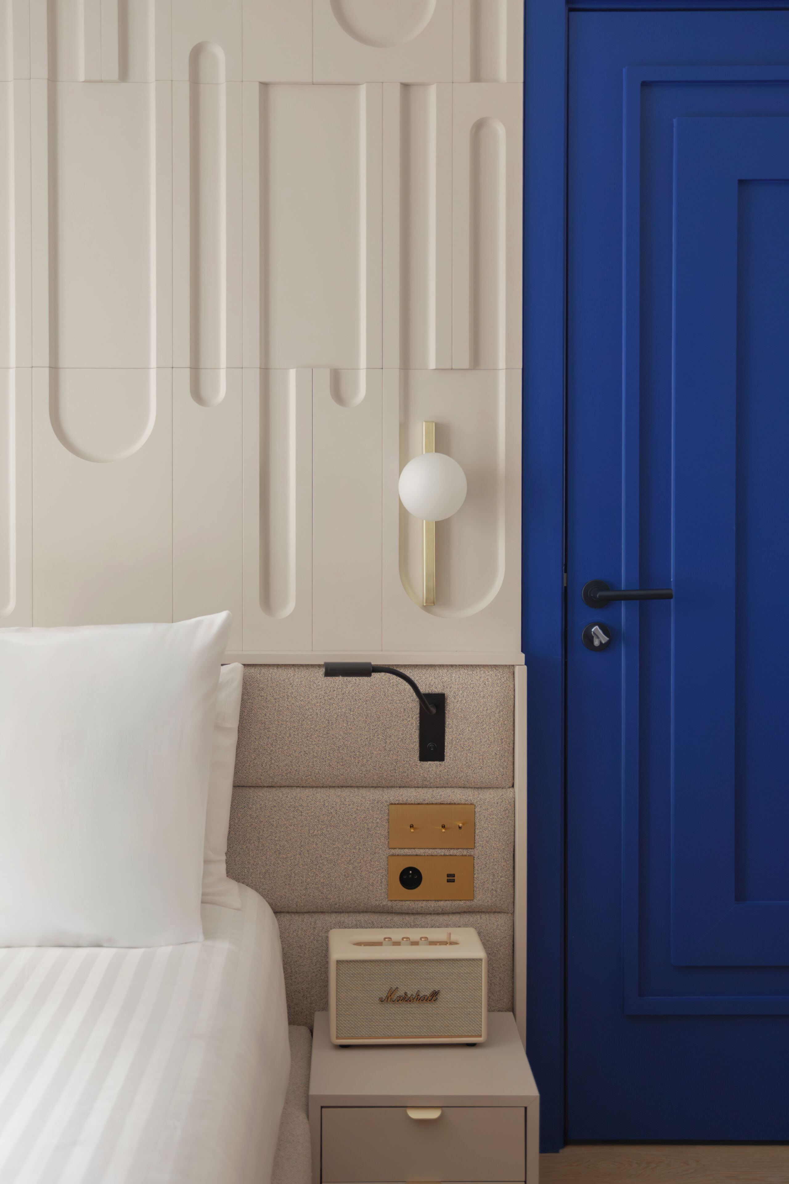 A bedroom with a bed, a nightstand with a radio, a blue door, and a wall with geometric patterns.