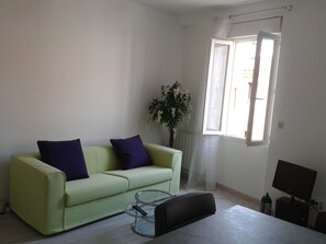 A living room with a green sofa, two purple cushions, a glass coffee table, a black ottoman, a potted plant, a window with white curtains, and a television on a stand. - A vacation in the sun and by the sea, with an apartment on the 1st or 2nd floor! (Elne)